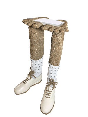 A sculpture from the 'A Wrinkle in the Binky' installation. The base is made of the two white shoes with sand laces that were in the Every Worm Deserves a Mansion video and mutant dew sculpture. The feet are flat on the ground and have sand legs coming out of them that end at the knee. Instead of knees there is a flat layer like a table shape, but the table top is shaped like two feet and covered in sand. There is also an indentation on the top of these flat sand feet that holds a white computer trackpad. The legs have white sweat socks with tin metal diamonds decorating them.